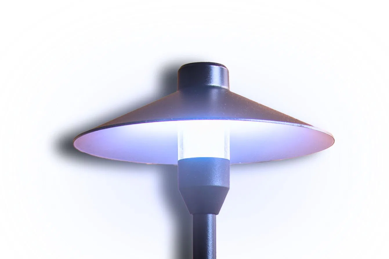 A modern outdoor lamp post with a wide, flat, circular shade and a cylindrical light in the center, glowing with a soft white light against a white background.