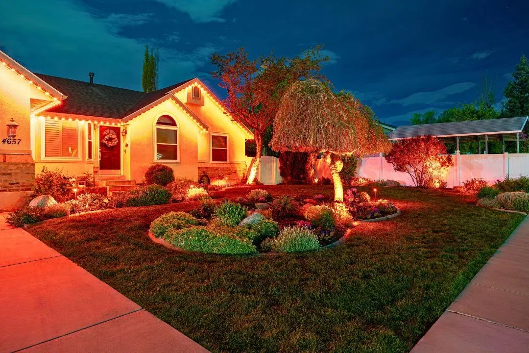 A single-story house with a well-manicured front yard is lit by orange string lights outlining the roof and trees, creating a warm, inviting glow at night. Shrubs, rocks, and a curved walkway enhance the landscaping.