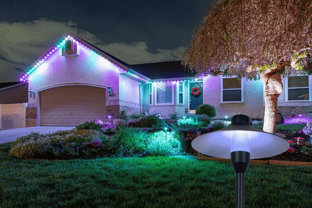 A house at night is decorated with colorful lights along the roof and garden. A bright outdoor solar lamp is illuminated in the foreground near a tree and landscaped yard.