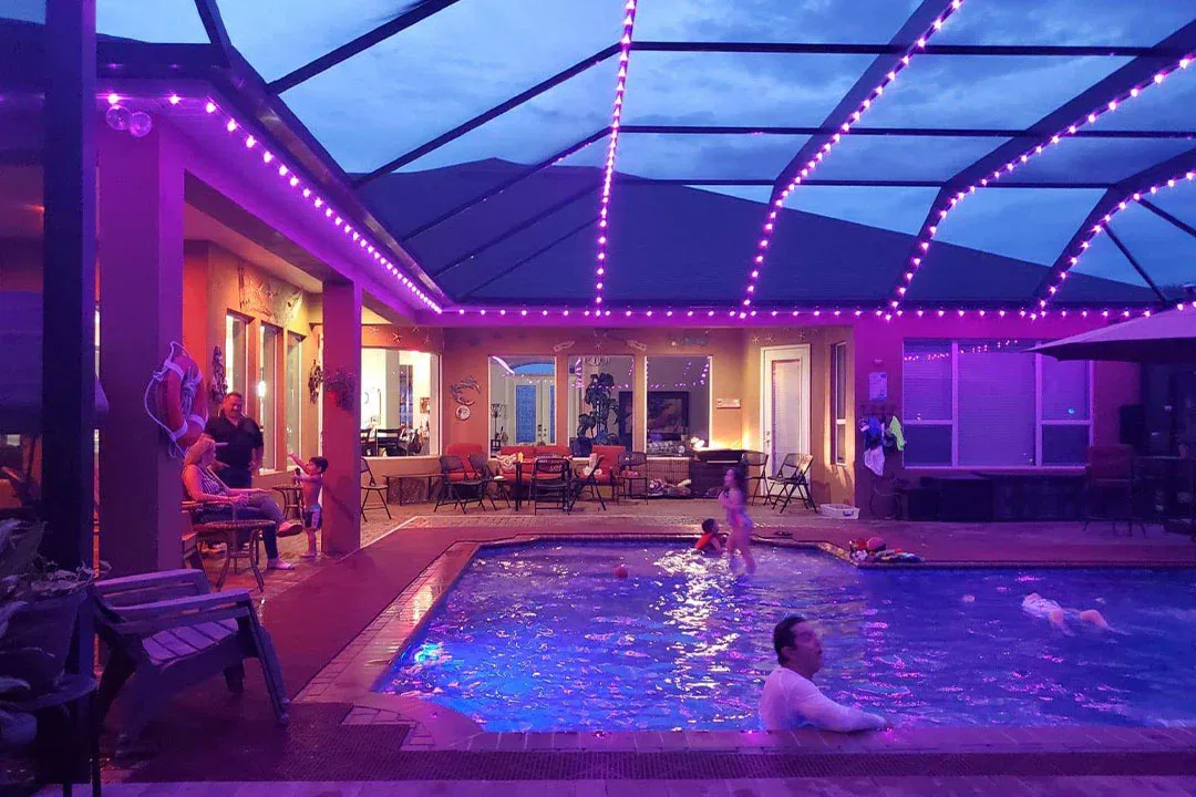 People enjoy an evening at a screened-in pool area, illuminated by purple string lights. Some swim in the pool while others sit and talk near tables and chairs under a covered patio. The sky is twilight blue.