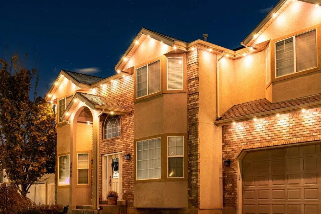 A two-story house with brick and stucco exterior is lit by warm, white lights outlining the roof and windows at night. A tree with autumn leaves stands to the left, and the sky is dark and starry.