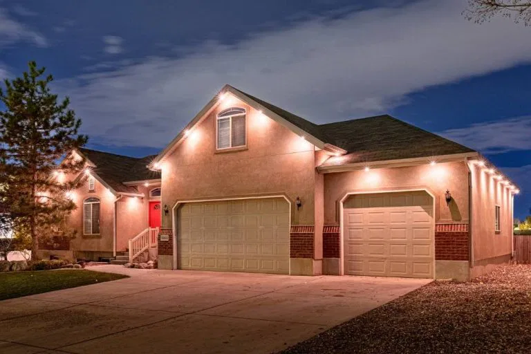 Single-story suburban house with beige walls, red brick accents, and a dark roof, illuminated by exterior lights at night. The home features a red front door, attached garages, and a concrete driveway.