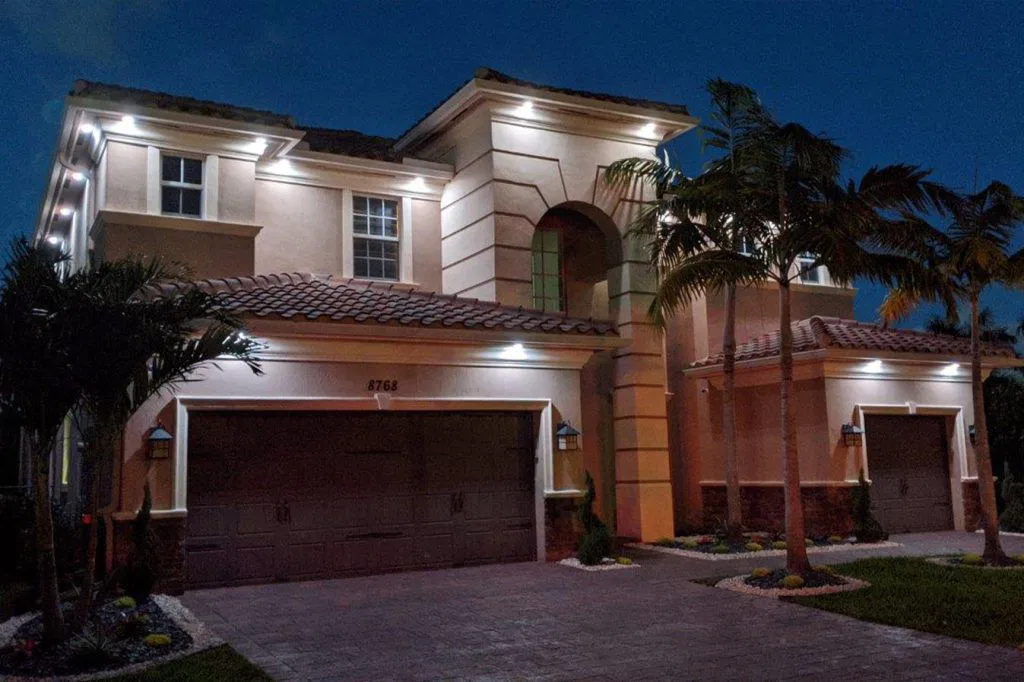 Two-story modern house at night with exterior lights illuminating beige walls, brown tiled roof, double garage doors, arched window, palm trees, and landscaped yard.