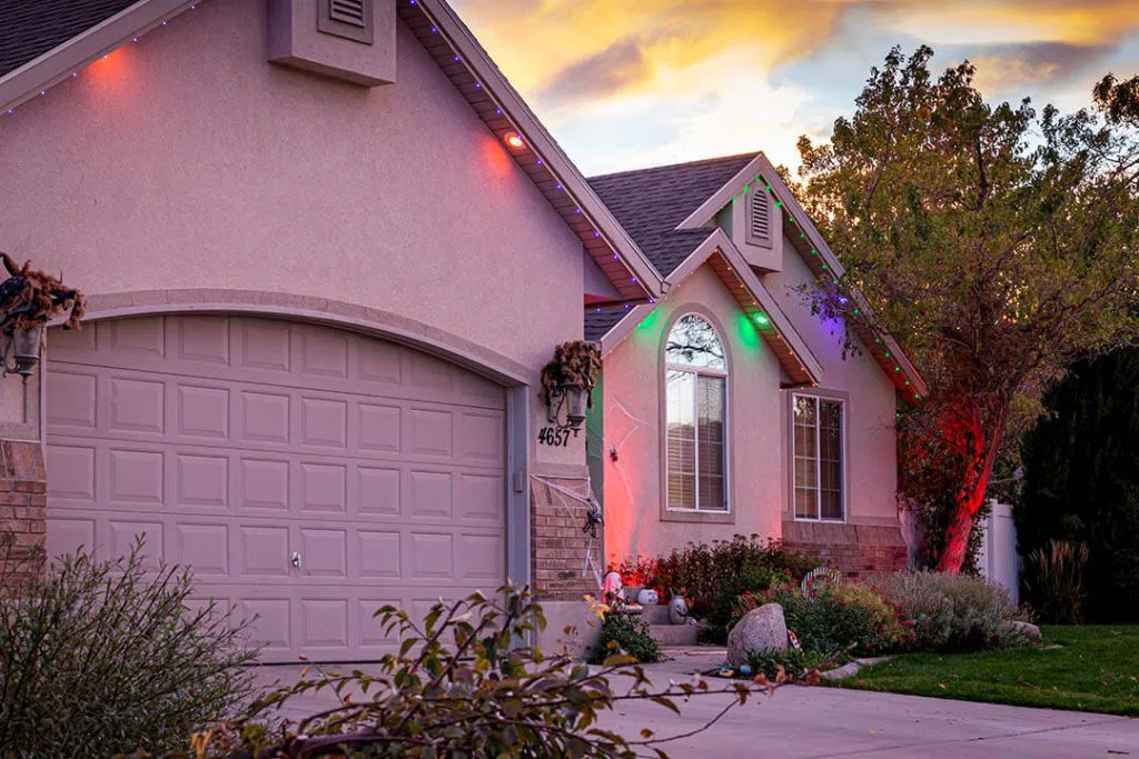 A suburban house at sunset with colorful holiday lights along the roof, a two-car garage, and a small front yard with bushes and trees. The sky is partly cloudy with warm colors.