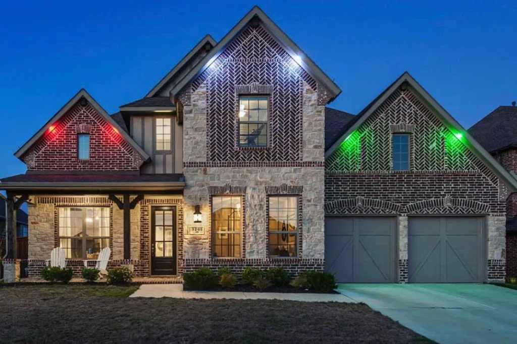 A two-story brick house with stone accents, illuminated by porch and exterior lights. The garage has two doors, and colored lights (red, white, green) highlight the gables. A small porch with chairs is visible on the left.