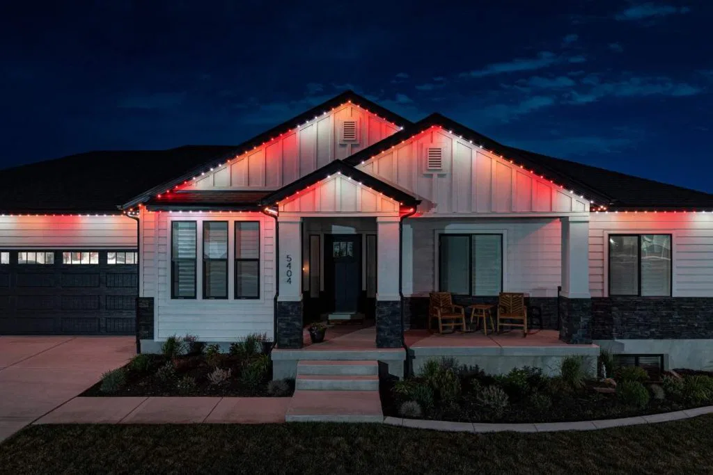 A modern single-story house at night with red and white lights outlining the roof, a dark garage door, two chairs on the porch, and well-kept landscaping in front.