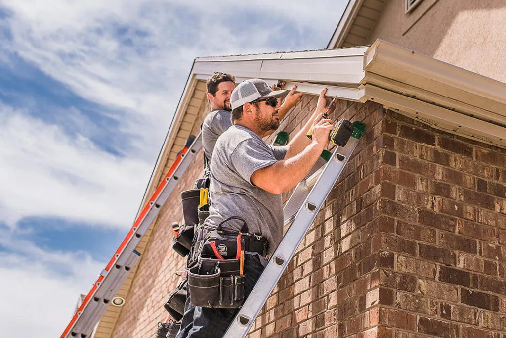 Two men wearing tool belts stand on ladders and work on the gutter of a brick house under a blue sky with clouds.