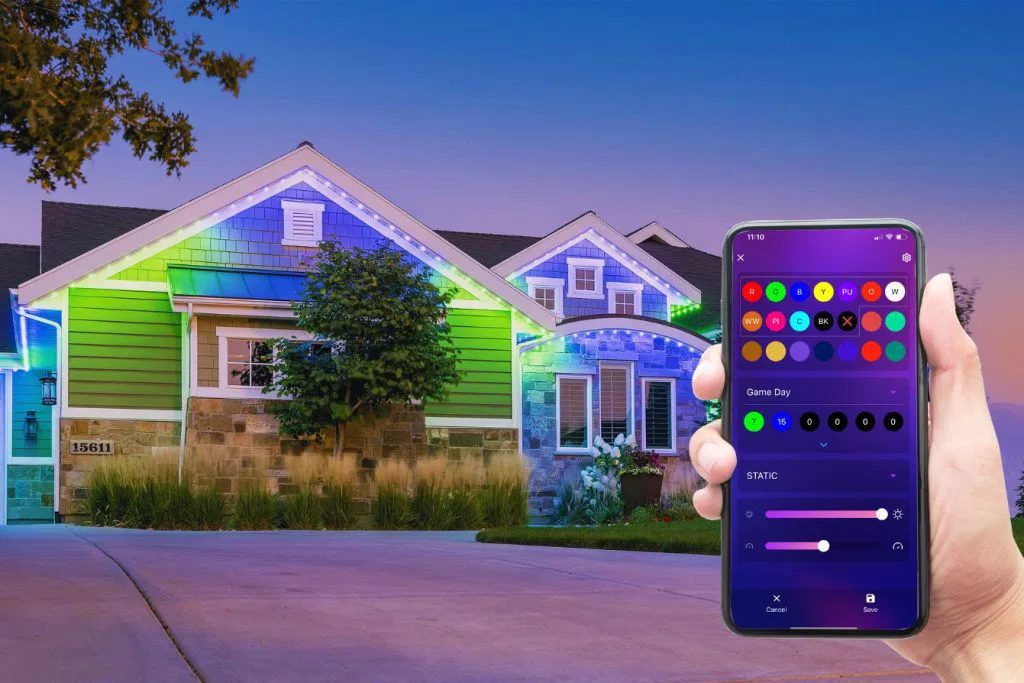 A hand holds a smartphone with an app controlling colorful LED lights on the exterior of a modern house at dusk. The house is decorated with green, blue, and white lights along the roofline.