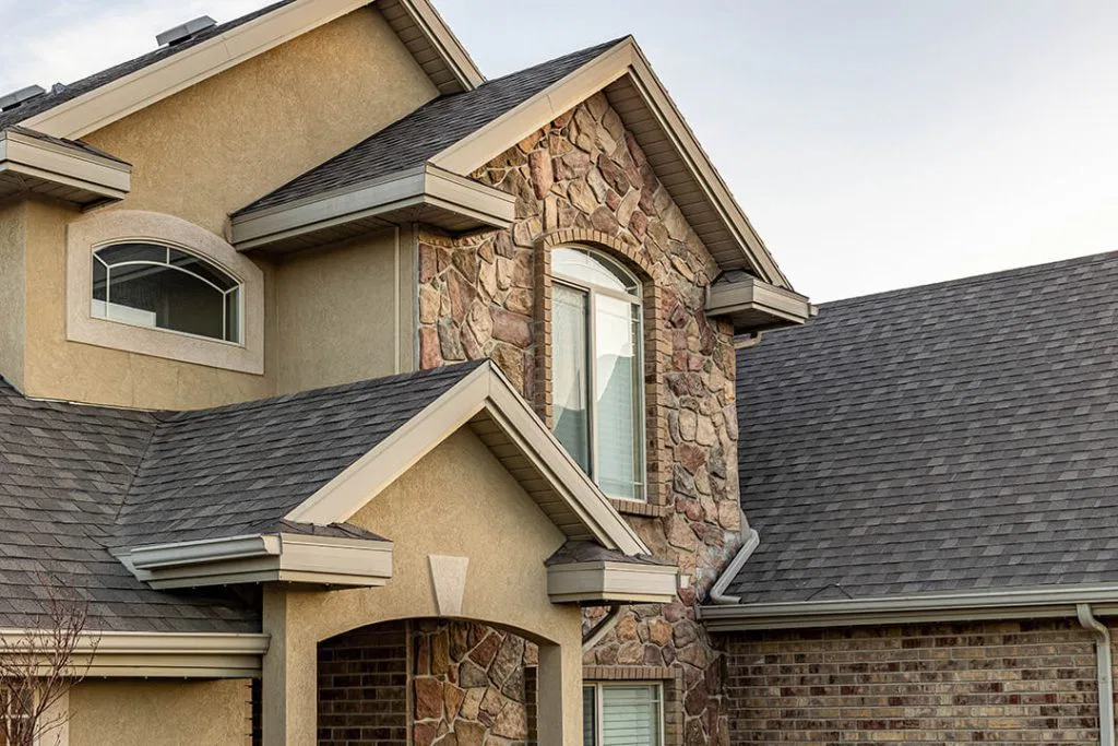 Two-story house exterior with beige stucco, decorative stone accents, arched and rectangular windows, and a gray shingle roof. The sky is clear in the background.