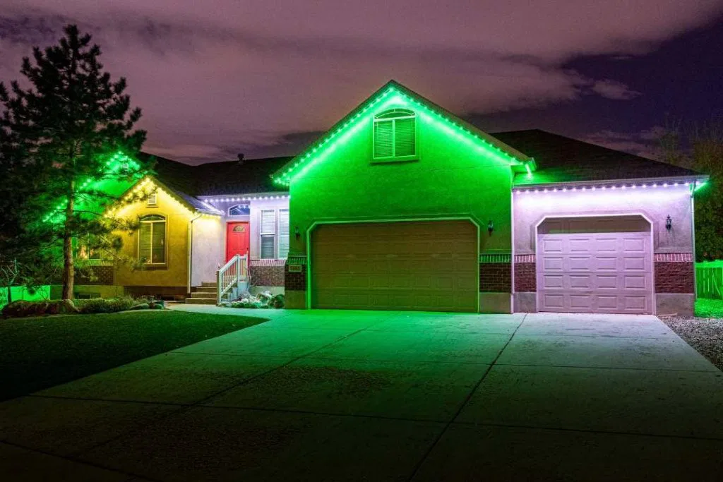 A single-story house at night is lit by green and white LED lights outlining the roof, with a tree on the left and a wide driveway in front. The sky is cloudy with a purple tint.