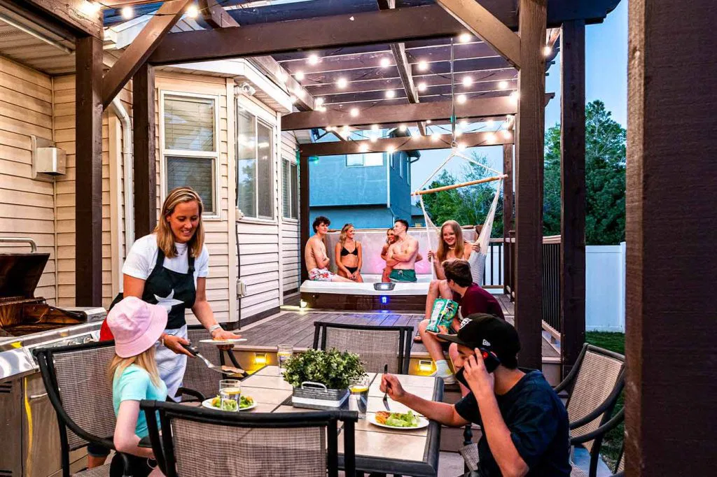 A woman serves food at a patio table where two kids are seated, while five people relax and talk in a raised hot tub under string lights in a backyard setting.