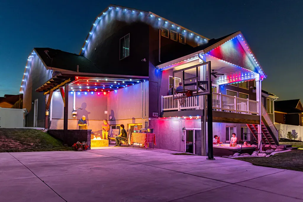 A two-story house at night decorated with colorful string lights, with people sitting and relaxing on a patio and balcony. A basketball hoop stands on the driveway in the foreground.