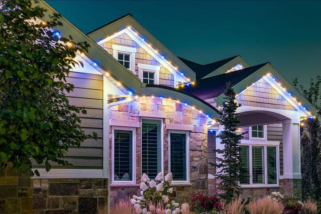 A modern house at dusk with stone and siding exterior, large windows, a front porch, and colorful string lights outlining the roof. Landscaping includes trees, shrubs, and white flowers in the garden.