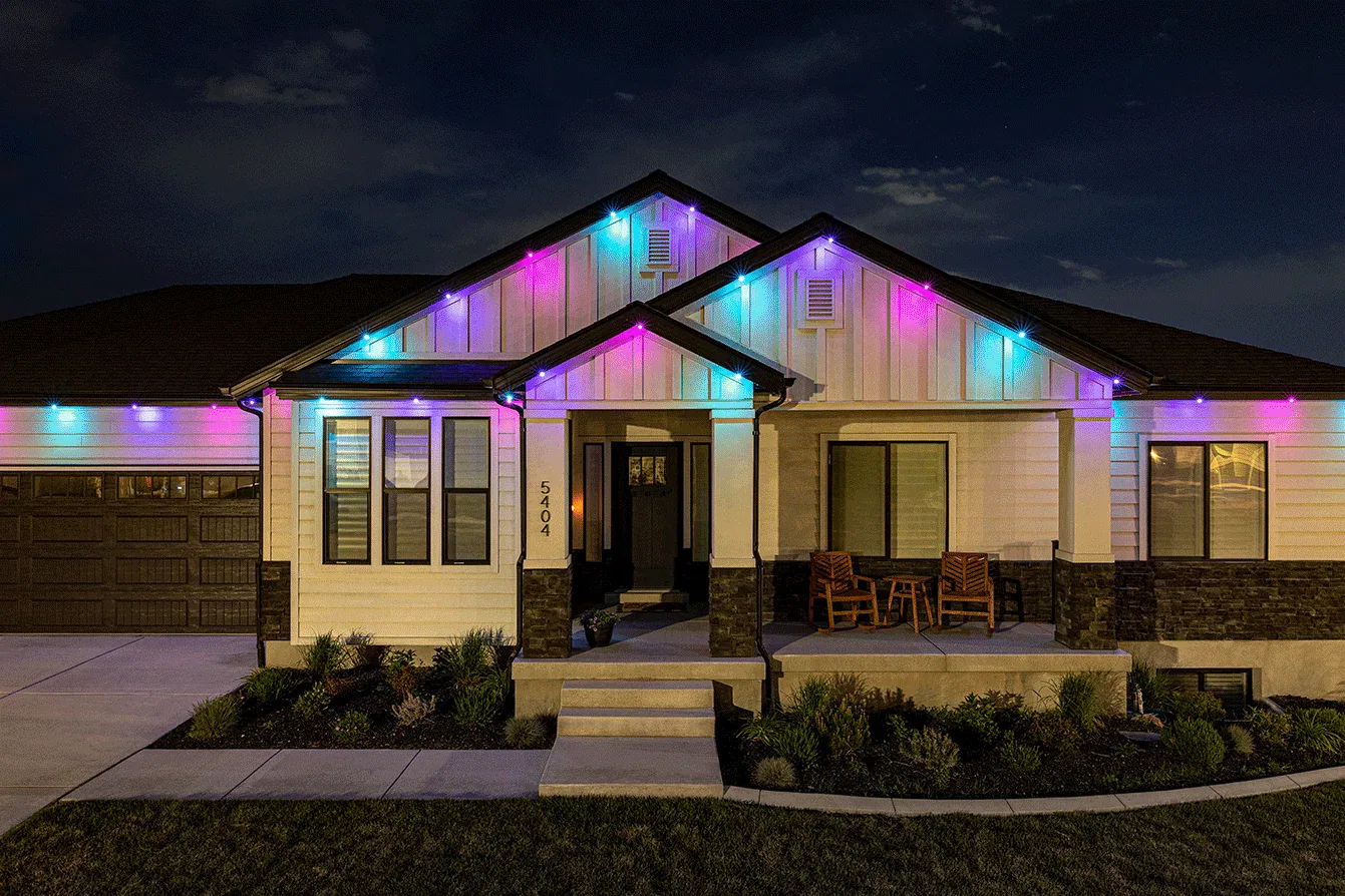 A modern single-story house at night, with colorful string lights in blue, purple, and pink decorating the roofline. The front porch has two wooden chairs, and the garden is neatly landscaped.