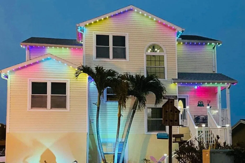 A two-story house decorated with multicolored string lights along the roof at dusk, with palm trees and a birdhouse in the front yard.