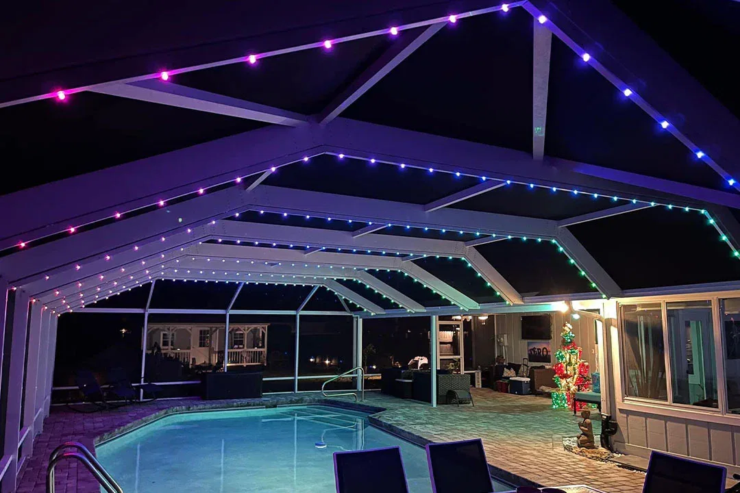 A screened-in pool area at night is decorated with colorful LED strip lights along the ceiling. Lounge chairs and patio furniture surround the pool, and a Christmas tree with lights stands near the entrance.