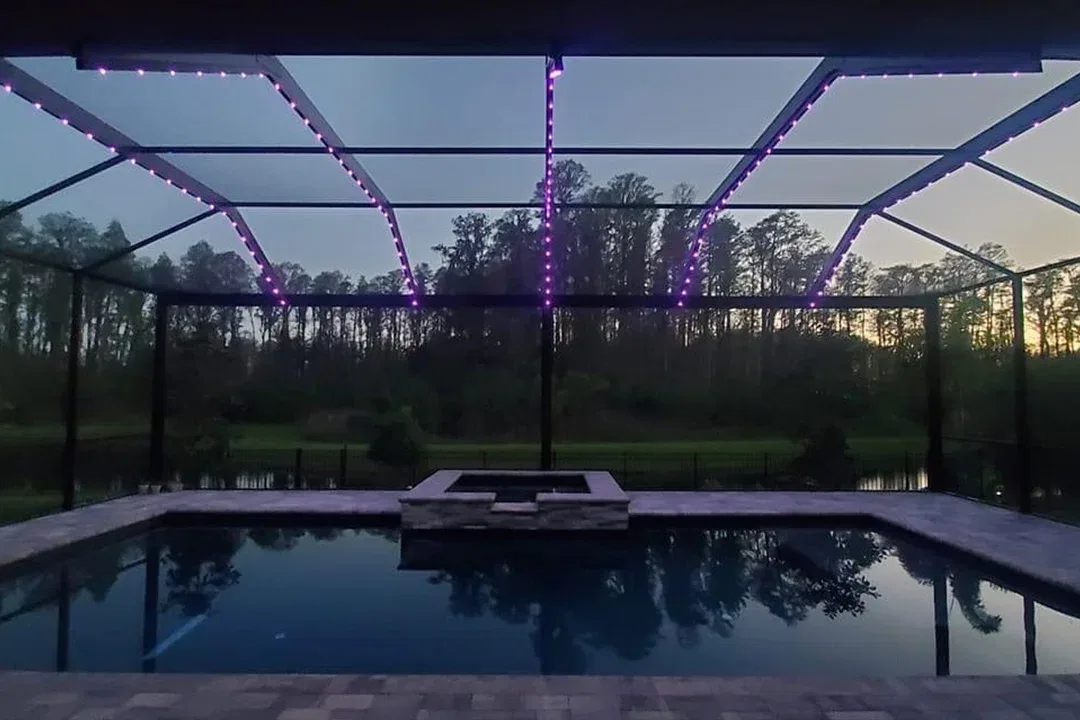 A screened-in pool and hot tub at dusk, with purple string lights overhead and tall trees reflected in the water, set against a backdrop of woods and a fading sunset.