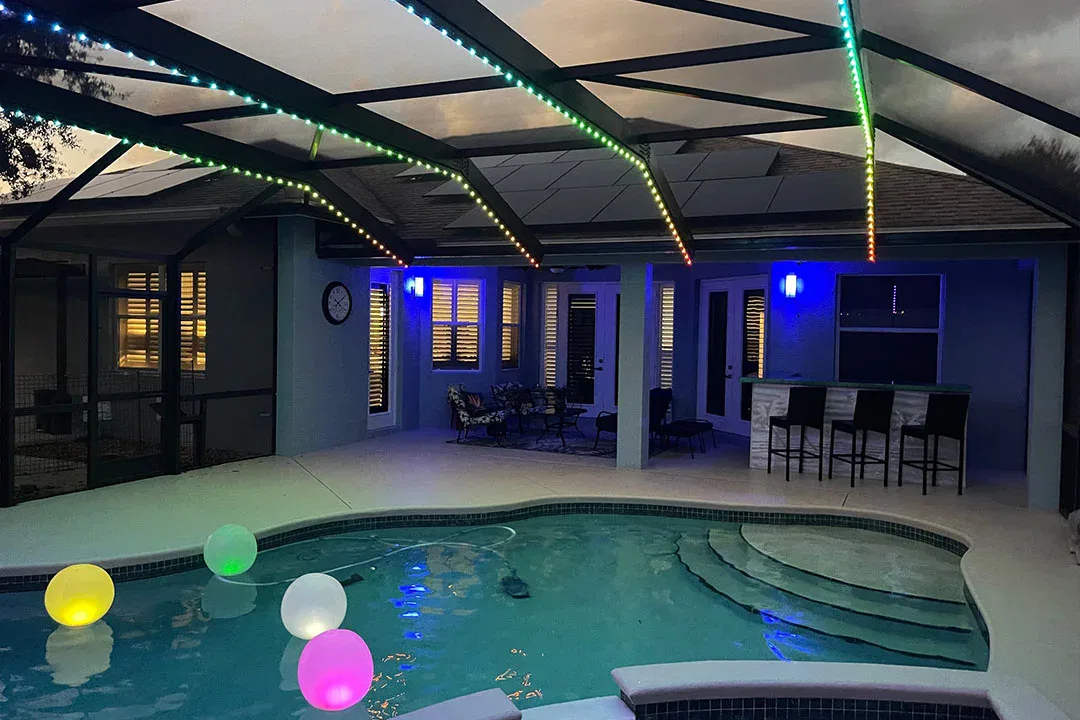 A backyard pool at dusk features glowing, colorful balloons floating in the water. LED strip lights and wall lights illuminate the covered patio area, which includes seating and a bar with chairs.