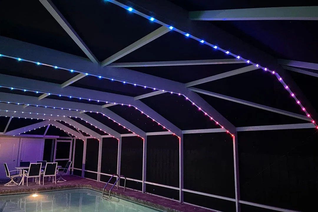 A screened-in patio with a pool features colorful LED strip lights along the ceiling beams, glowing blue, purple, and red. Patio furniture is arranged near the pool under the illuminated structure.