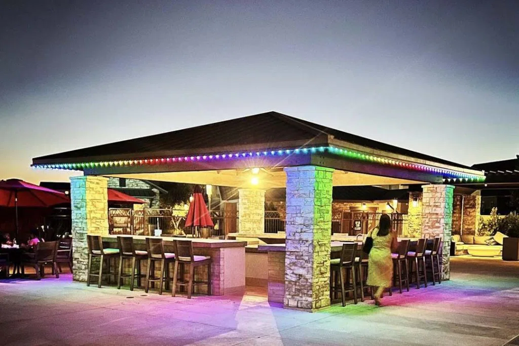 An outdoor bar with stone pillars and a dark roof is lit by multicolored string lights at dusk. Barstools surround the counter, and a person is walking nearby. The sky is a gradient of deep blue and yellow.