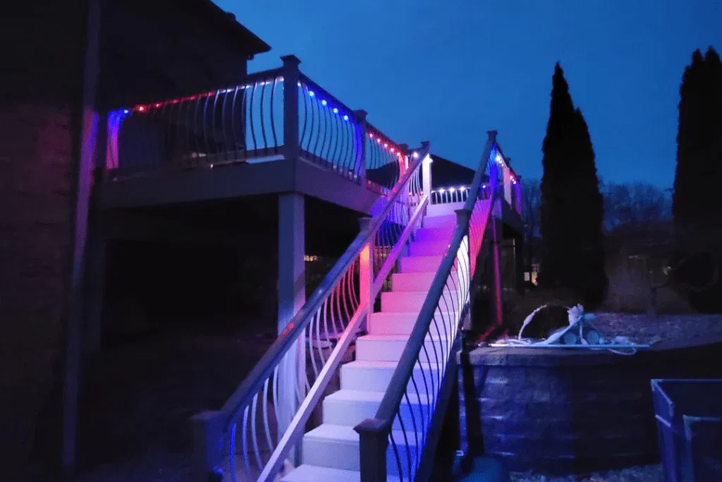 A staircase leading to an upper deck is illuminated with red, white, and blue lights at night. The decks railing is also decorated with matching string lights. Trees and a dark sky are visible in the background.