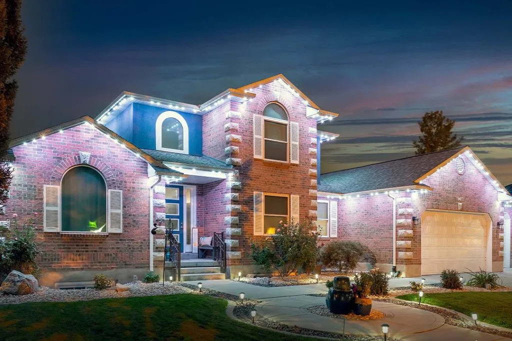 Two-story brick house with arched windows and white trim, illuminated by white string lights at dusk. The front yard has shrubs, rocks, and pathway lights leading to the entry and garage.