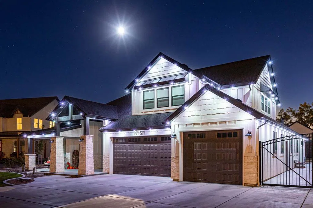 A modern two-story house is illuminated with white string lights along the roofline at night. The driveway and garage are visible, with a bright full moon shining in the clear sky above.