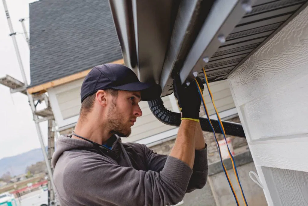 A man wearing a cap and gloves installs or repairs a gutter on the roof edge of a house, using tools and guiding wires. The house exterior and nearby outdoor scenery are visible.