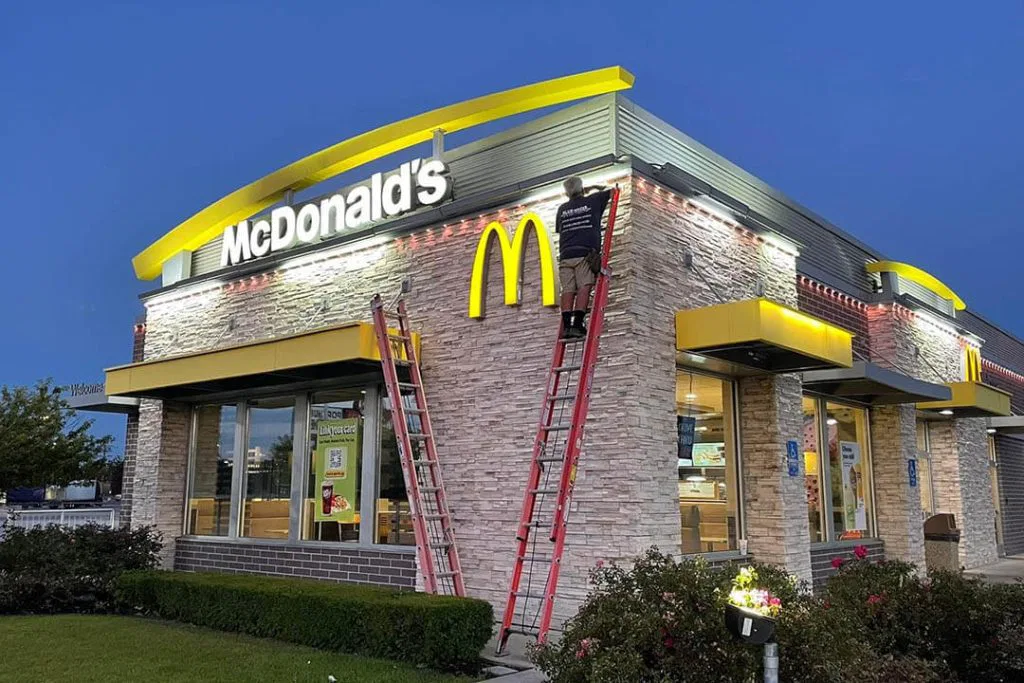 A worker stands on a ladder installing or repairing lights on the exterior of a McDonalds restaurant at dusk. The building features stone walls, large windows, and prominent yellow arches.