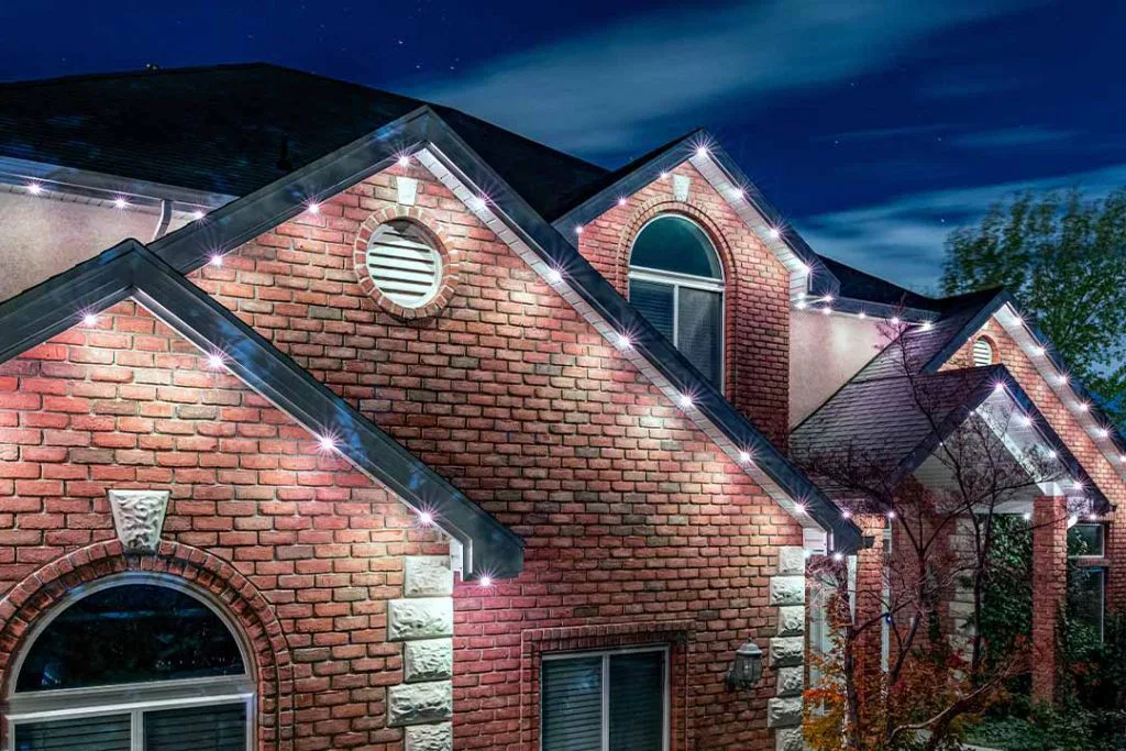 A brick house at night with white string lights outlining the roof edges, creating a festive and welcoming appearance under a dark, starry sky.
