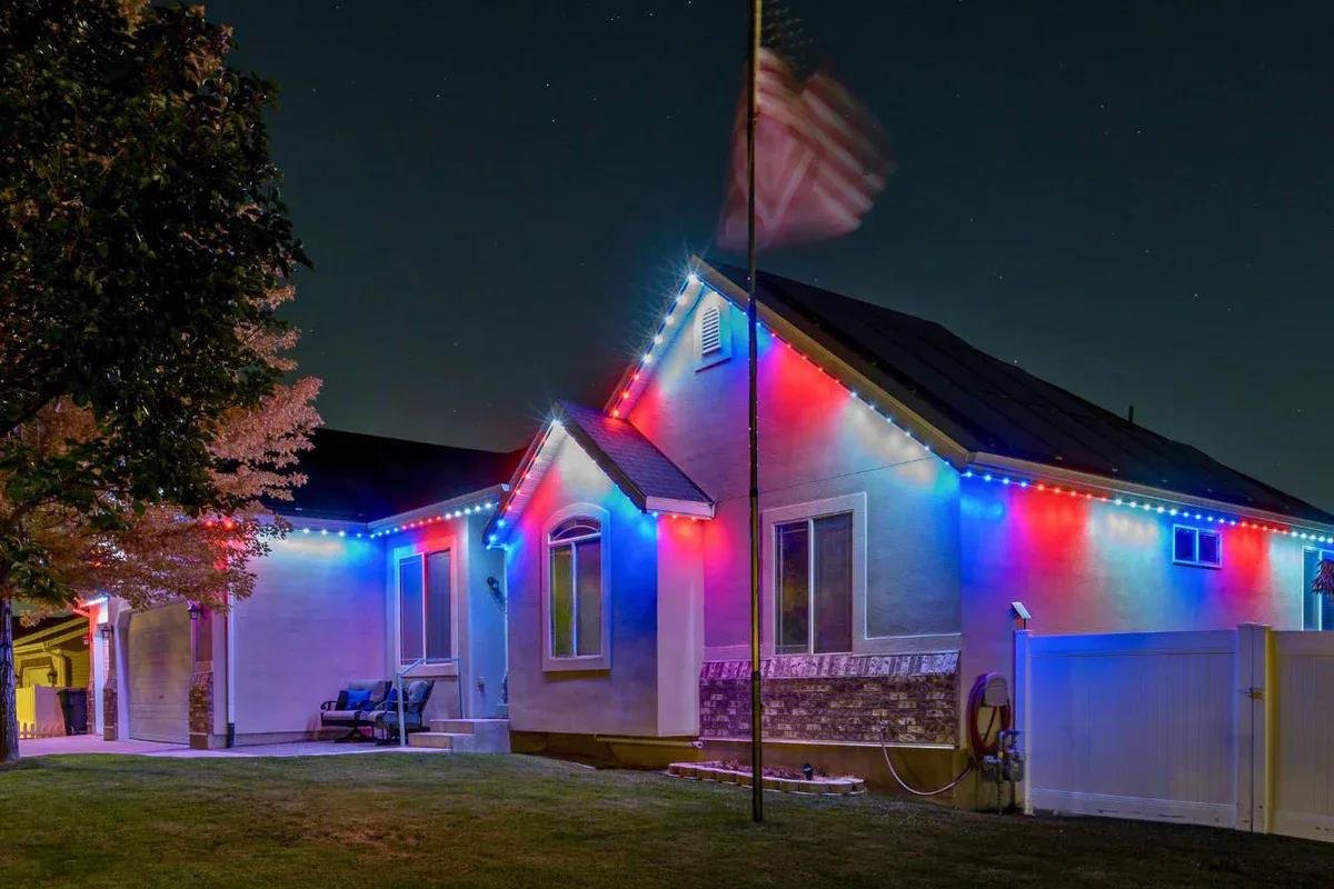 A house at night is decorated with red, white, and blue lights along the roofline. An American flag waves on a pole in the front yard, and a large tree is partially visible on the left.