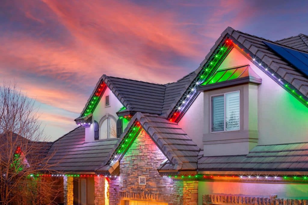 A house with stone accents and a tiled roof is decorated with red, green, and white holiday lights along the roofline. The sky is colorful with dramatic pink and orange clouds at sunset.