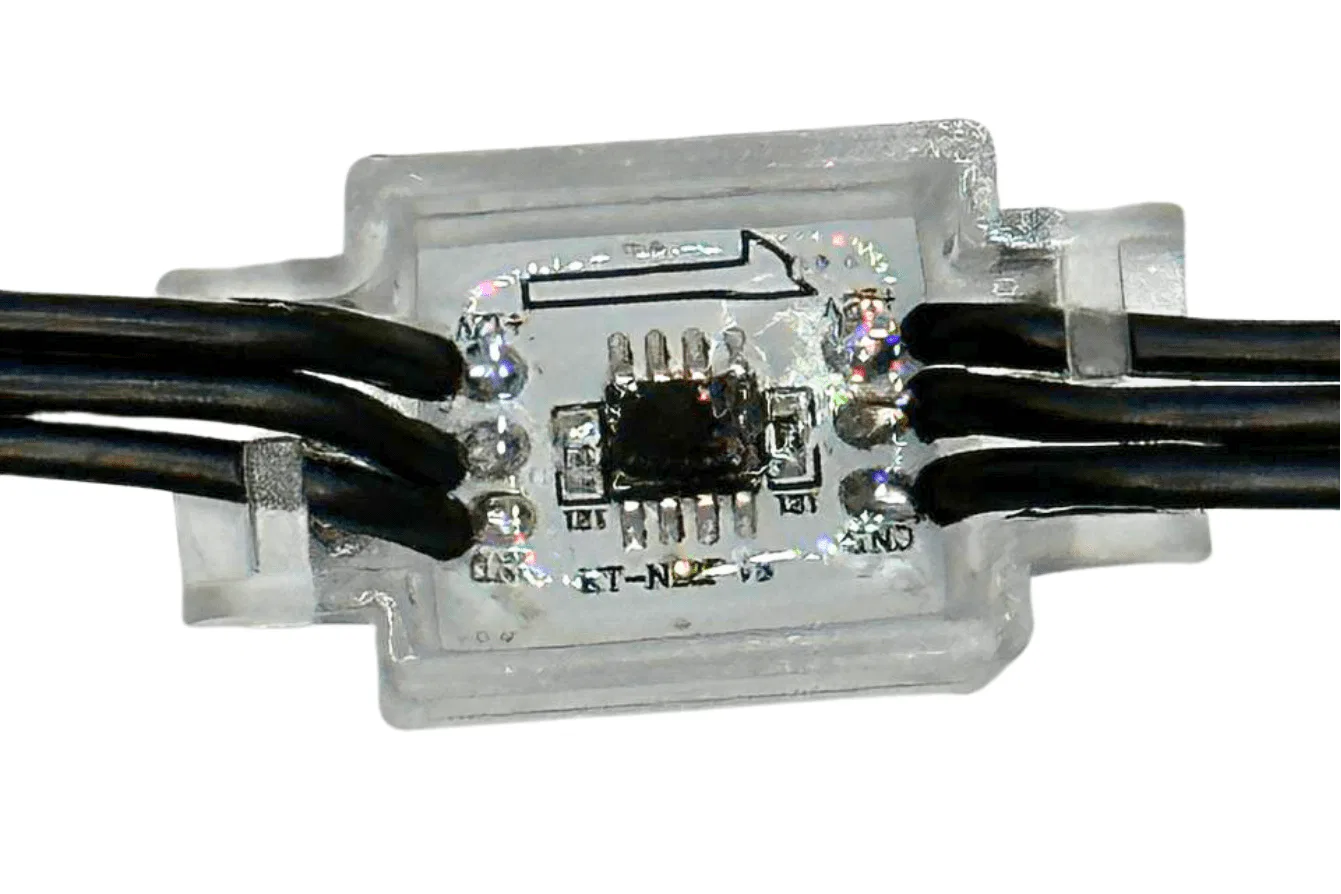 Close-up of a small electronic circuit board encased in transparent plastic, with black wires connected to each side and an arrow printed on the casing. The circuit board has several visible components and connections.