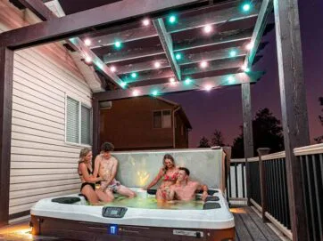 Four people in swimsuits relax in a hot tub under a pergola with string lights at dusk, on a deck attached to a house.