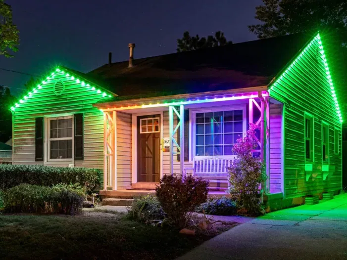 A single-story house at night is decorated with vibrant, multicolored LED lights along the roofline and porch, creating a bright and festive glow against the dark sky. Shrubs and a white porch swing are visible in front.
