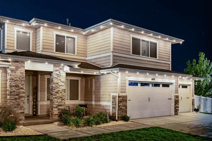 Two-story suburban house at night with stone accents, beige siding, white trim, and exterior lights along the roofline. There is a double garage, a small landscaped yard, and a white fence with trees in the background.