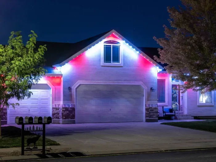 A house at night with white, blue, and red lights decorating the roofline. The garage doors are closed, and trees are visible on either side of the driveway, which is empty. Three mailboxes stand at the curb.