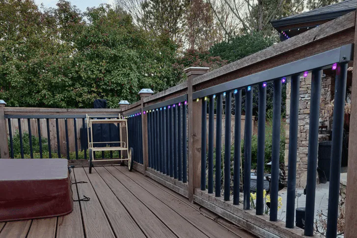 A wooden deck with black railings decorated with multicolored string lights, a closed hot tub on the left, a folded chair, and lush green trees in the background.