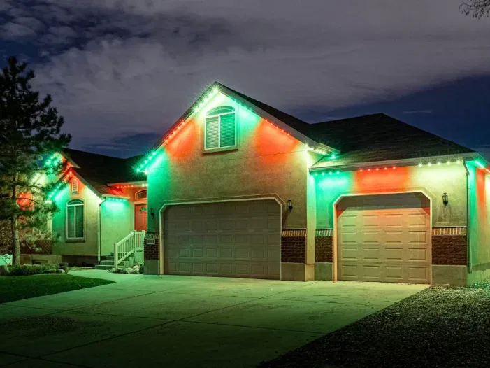 A house at night with red and green lights outlining the roof and garage, creating a festive appearance. The sky is partly cloudy, and the driveway and yard are visible in the foreground.