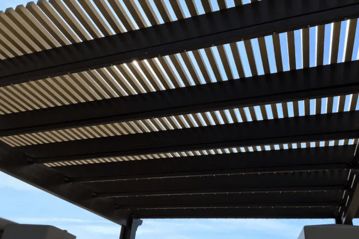 A close-up view from below of a pergola with evenly spaced horizontal slats, casting striped shadows as sunlight filters through. The sky is clear and blue in the background.