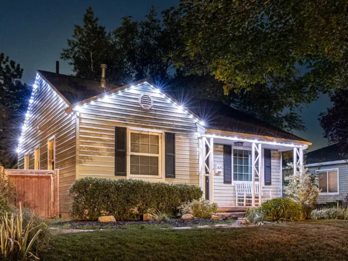 A small house at night with white string lights outlining the roof, a front porch with a white rocking chair, shrubs in front, and trees surrounding the yard.