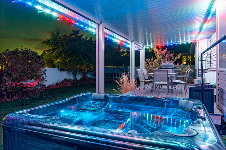 Covered patio at night with colorful LED string lights, a glowing hot tub in the foreground, outdoor dining table and chairs, and a hammock on a grassy lawn surrounded by bushes and trees.