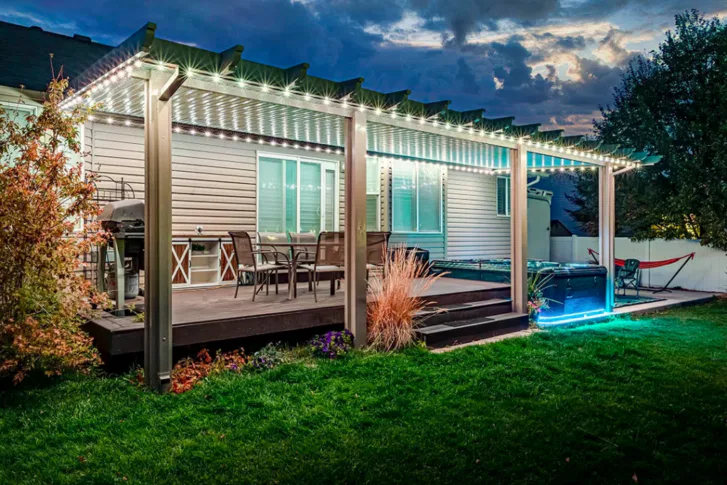 A backyard patio with a pergola decorated with string lights, featuring outdoor furniture and a hot tub, sits on a lawn at dusk. The house and yard are softly illuminated, creating a cozy atmosphere.