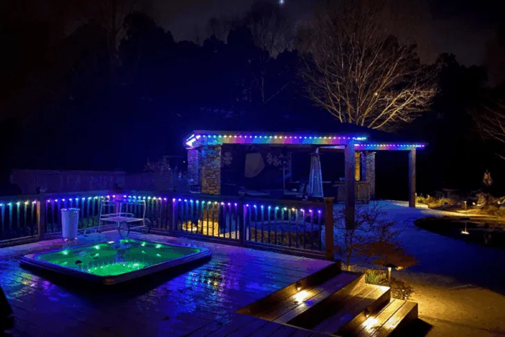 A backyard at night with colorful LED lights outlining a pergola and deck, a hot tub glowing green, and trees and a pool in the background, creating a vibrant, cozy atmosphere.