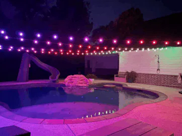 A backyard pool at night, lit by purple and pink string lights. Theres a waterslide, a small rock waterfall, poolside seating, and a white house wall in the background. The area glows with colorful reflections.