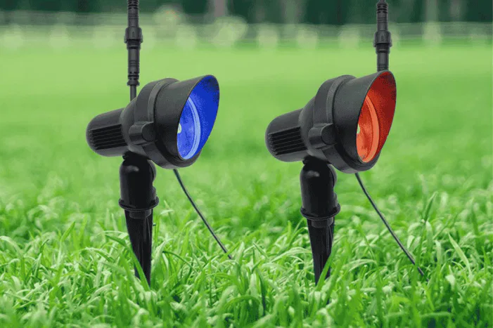 Two black outdoor spotlights are staked in green grass. One light glows blue and the other glows red, with blurred fencing and greenery in the background.