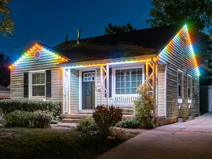 A small house at night with colorful LED lights outlining the roof and front porch, creating a festive appearance. Shrubs and a small garden line the walkway leading to the front door.