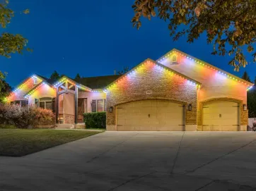 A single-story house with three garages is lit by multicolored string lights along the roofline at night, surrounded by trees and a well-kept lawn.