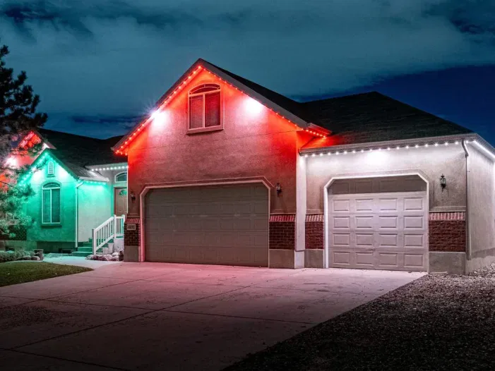 A suburban house at night with its roofline decorated with red, white, and green lights. The driveway and garage are visible, and the sky is partly cloudy. Another house with green lights is partially visible on the left.