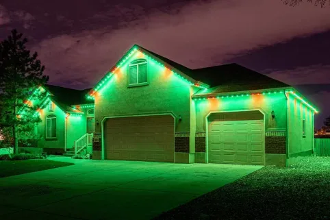 A single-story house at night with green and red lights outlining the roof, garage doors, and front porch. The driveway and yard are illuminated by the colored lights.